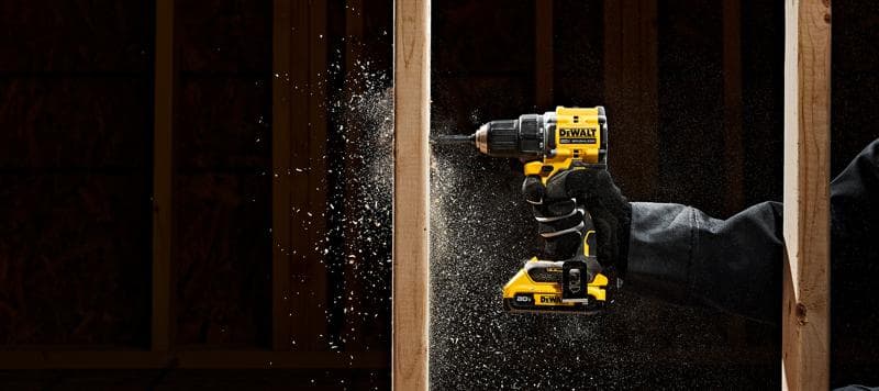A person wearing gloves uses a yellow and black DEWALT cordless drill to drill into a wooden beam, with wood particles flying from the drilling point.