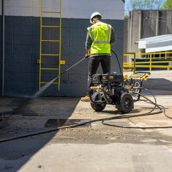 A person wearing a safety vest and helmet uses a DEWALT pressure washer on an outdoor concrete surface near a building with a yellow ladder.