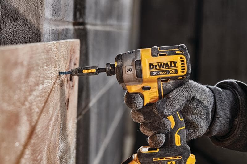 A person wearing work gloves uses a yellow and black DEWALT 20V MAX XR cordless impact driver to drive a screw into a wooden board.