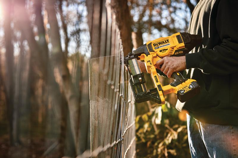 A person is using a yellow DEWALT cordless fencing stapler (model DCFS950B) to attach wire fencing to wooden posts outdoors.