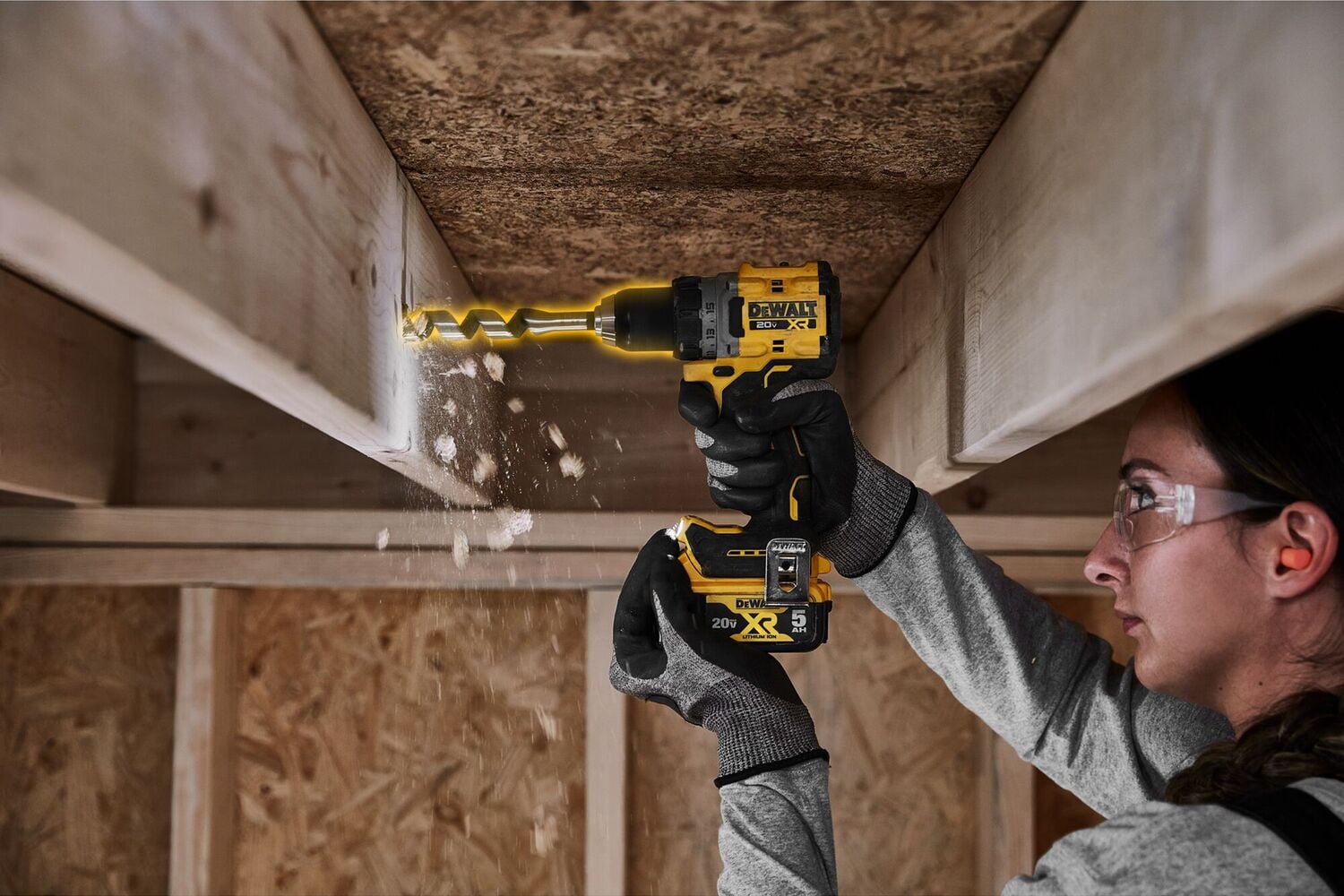 A person using a DEWALT cordless drill to bore a hole into a wooden beam, with wood shavings falling below. The person is wearing gloves and the setting appears to be a construction site.