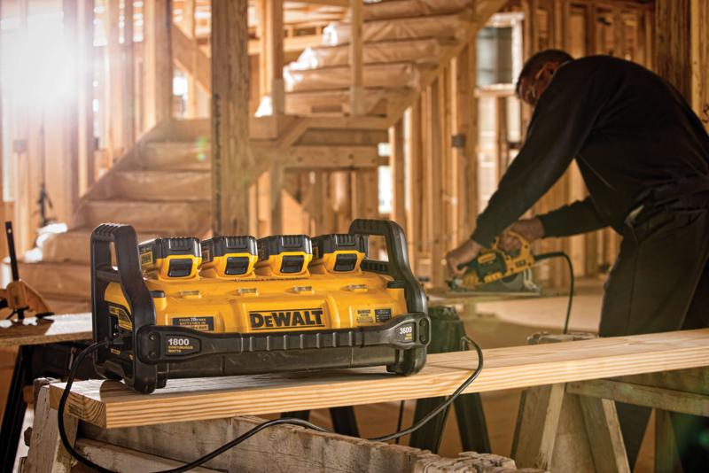A DEWALT portable power station model DCB1800B with four battery slots is placed on a wooden plank in a building under construction, with a person using a power tool in the background.
