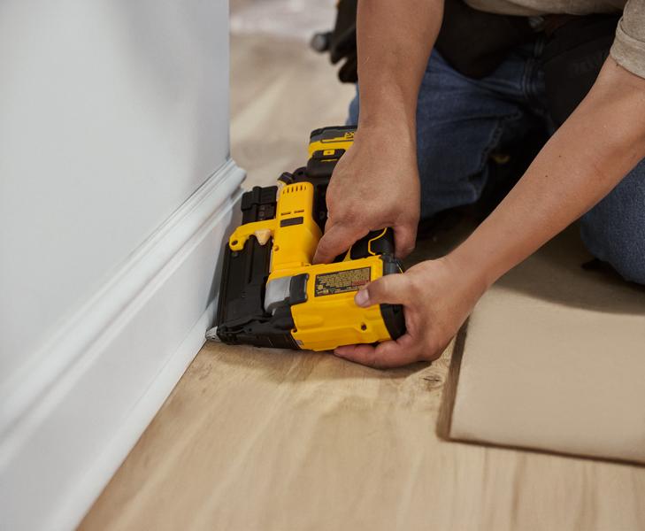 A person kneeling on the floor uses a yellow DEWALT cordless nailer to secure base molding to the wall in a home interior setting.