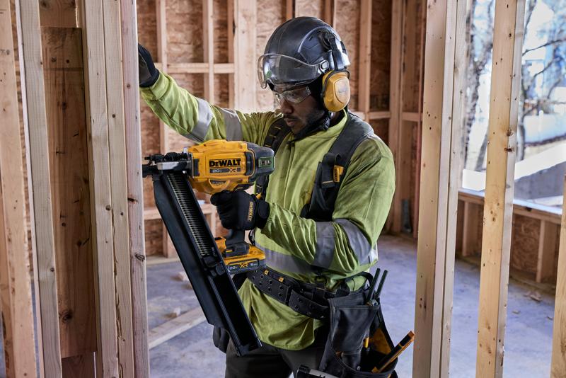A construction worker wearing safety gear is using a DEWALT DCN920B nailer to secure wooden framing inside a building under construction. The worker's face is blurred.
