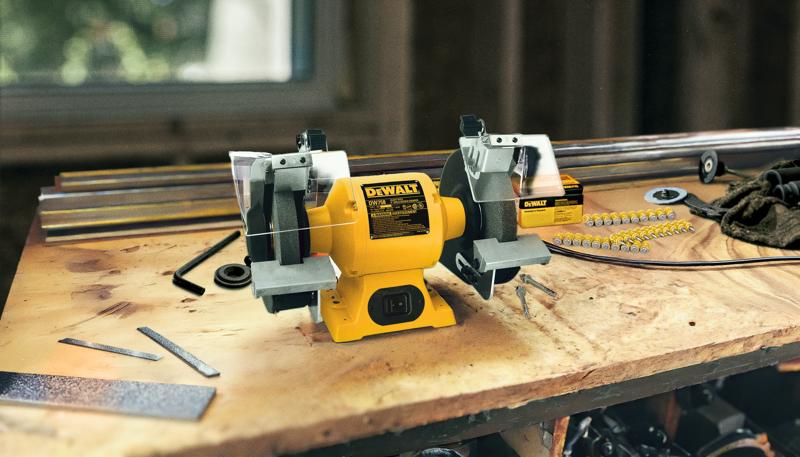 A DEWALT bench grinder (model DW758) is placed on a wooden workbench. The yellow and black power tool has two grinding wheels, one on each side, with protective guards and tool rests. Surrounding the grinder are various metal files, rods, and small workshop tools in a workshop environment.