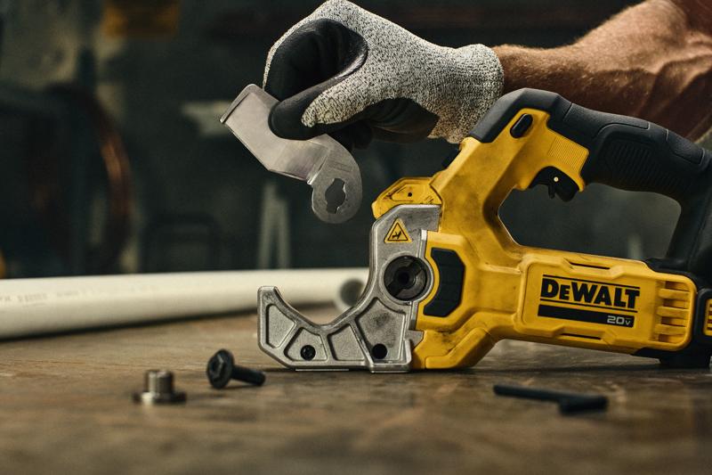A person wearing a protective glove is holding a blade attachment next to a yellow DEWALT 20V power tool, which is placed on a workbench. Small hardware pieces are visible on the table, and a white plastic pipe is in the background.
