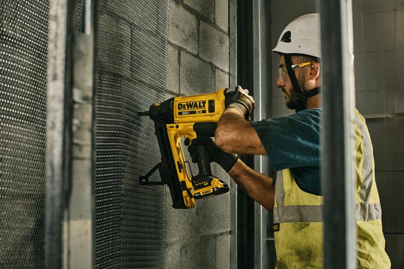 A construction worker wearing a safety vest and helmet uses a DEWALT cordless nail gun (model DGN845D1) to secure metal mesh onto a concrete wall inside a building.