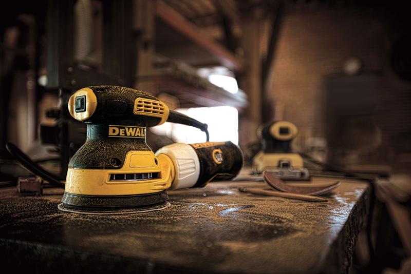 A DEWALT DWE6421 electric random orbit sander rests on a dusty workshop table, surrounded by sanding pads and tools, with another sander visible in the background.