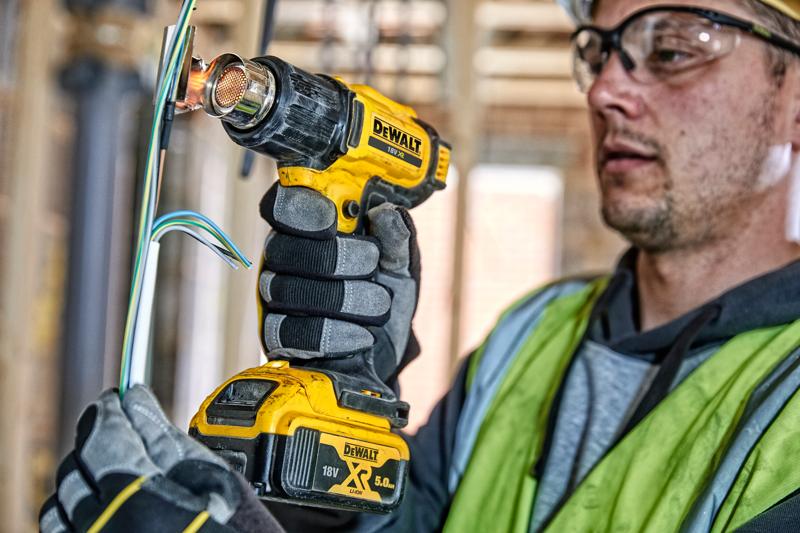 A person wearing gloves and a safety vest is using a DEWALT 18V XR cordless heat gun to work on electrical wires at a construction site. The person's face is blurred.