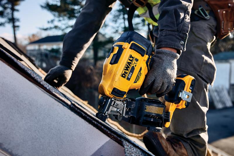 A worker using a yellow DEWALT cordless roofing nailer tool on a rooftop, wearing gloves and work clothes.