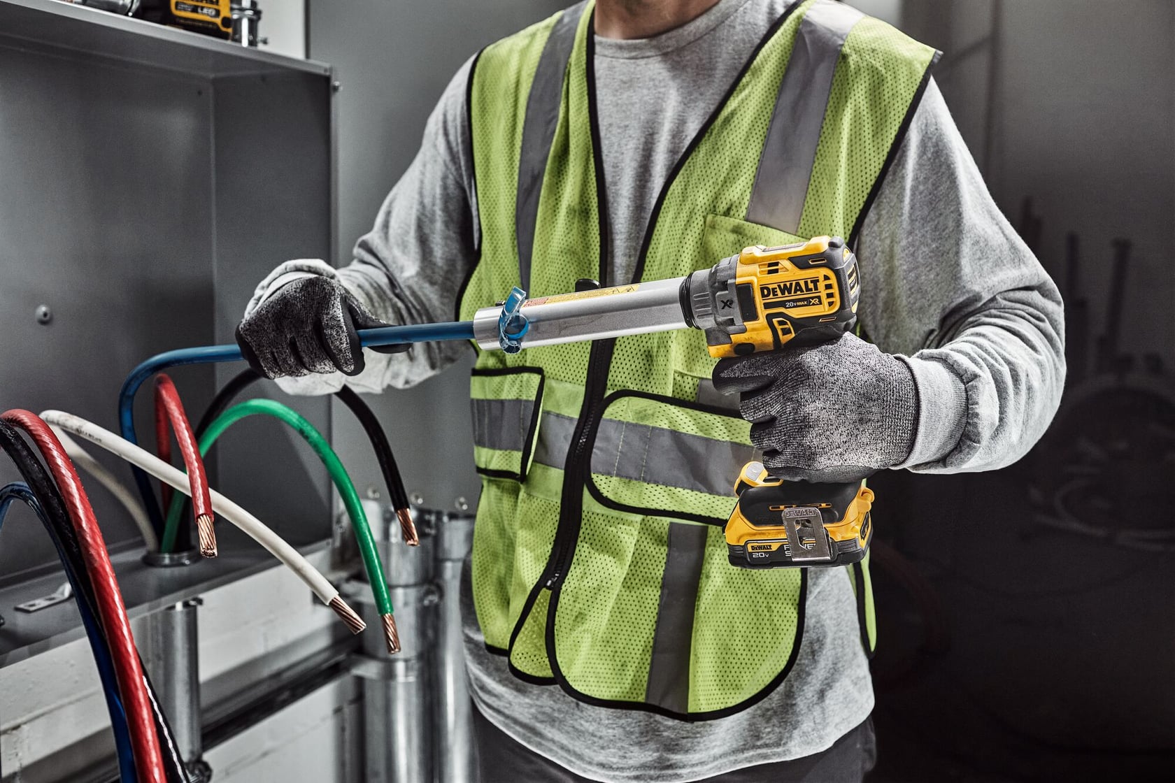 A person wearing a high-visibility safety vest and gloves is using a DEWALT cordless power tool to work with electrical cables in an industrial setting.
