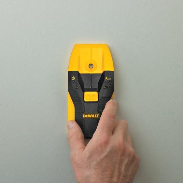 A hand holds a DEWALT electronic stud finder against a light-colored wall. The device is yellow and black, with a power button and AC detection indicator visible on the front.