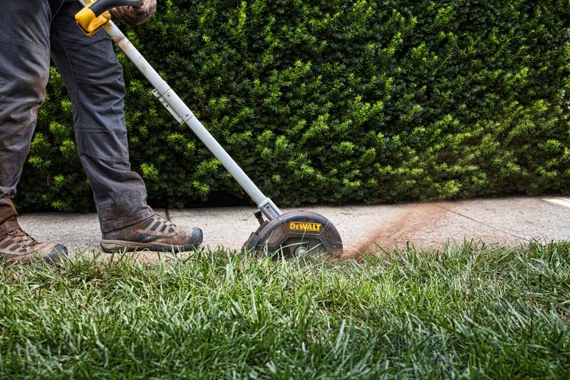 A person uses a DEWALT lawn edger to trim the edge of a grassy lawn next to a sidewalk, with green bushes in the background.