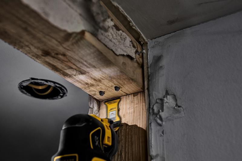 A DEWALT oscillating tool being used to cut wood in a corner of a construction site, with visible drywall and exposed timber.