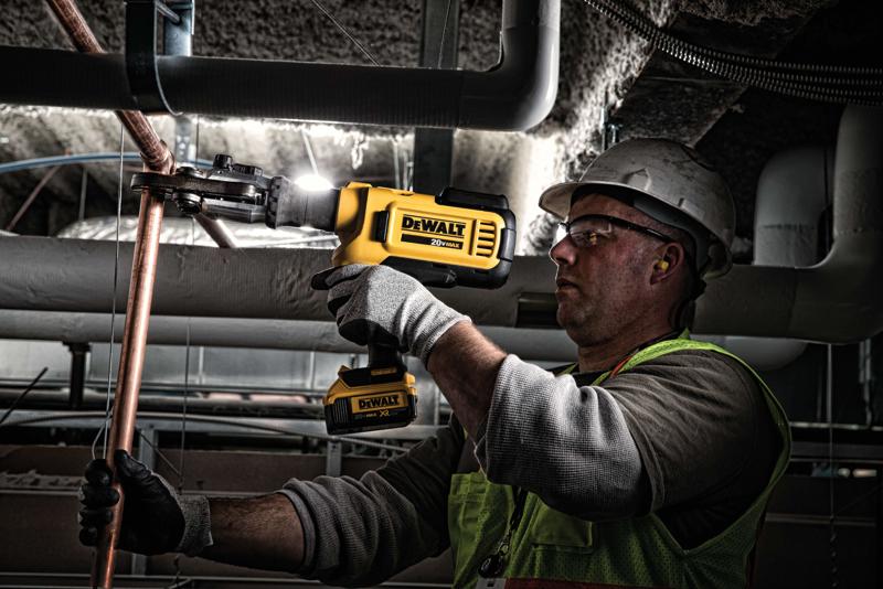A worker wearing gloves, a safety vest, and a hard hat uses a yellow DEWALT cordless tool to cut or crimp a copper pipe in an industrial or construction setting.