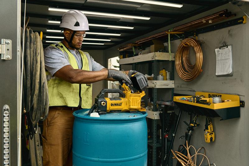 A person wearing a safety helmet and vest is using a DEWALT tool, model DCE050P1, on top of a blue barrel inside a workshop or storage area. The space has shelves, coiled copper tubing, and other tools and equipment visible.