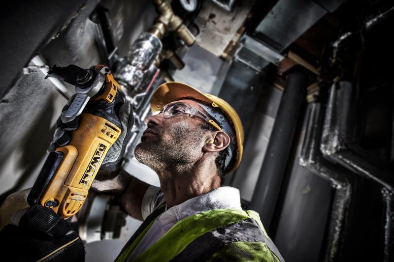 A worker in safety gear uses a DEWALT DWE315 power tool to cut into a wall in an industrial setting. The worker's face is blurred for privacy.