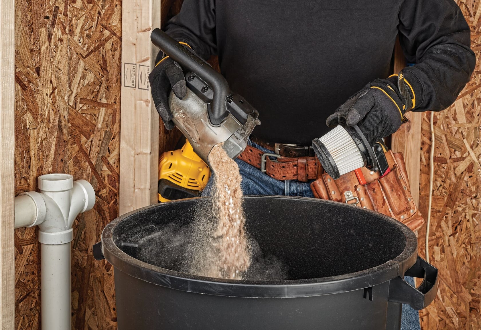 A person wearing gloves empties dust and debris from a DEWALT vacuum canister into a large black bin. The person is also holding a vacuum filter, and has a tool belt around their waist. The scene takes place in a workshop with wooden walls and plumbing pipes visible.