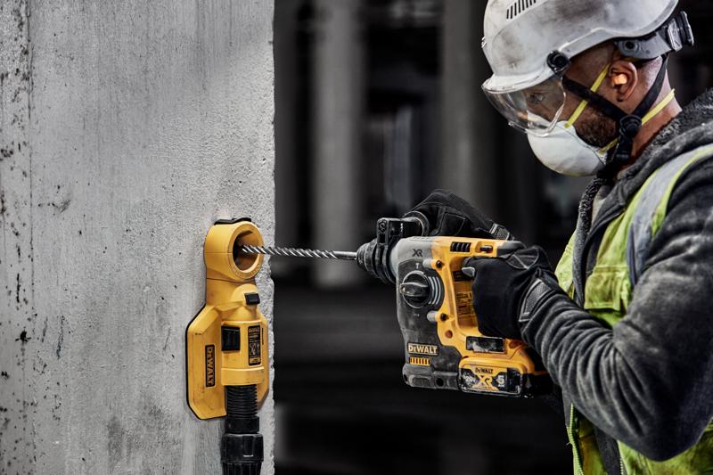 A construction worker wearing safety equipment is using a yellow DEWALT power drill to drill into a concrete wall. A DEWALT dust extraction attachment is connected to the wall to collect debris.