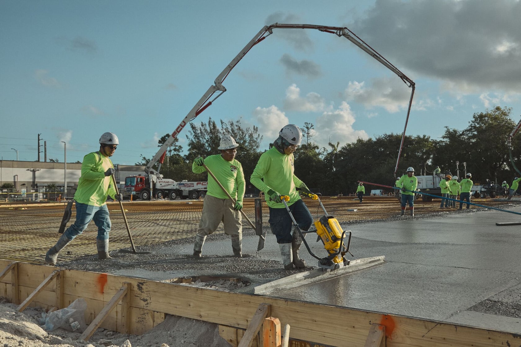 Construction workers wearing safety gear and green shirts are working on a concrete slab. One worker is using a DEWALT tool to smooth the freshly poured concrete, while others assist with the process. A concrete pump and machinery are visible in the background.