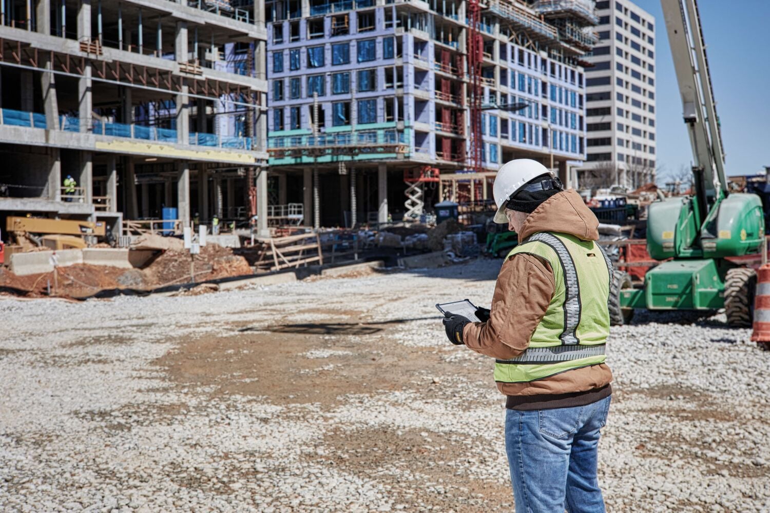 Person wearing a hard hat and safety vest using a tablet at a construction site with a partially built multi-story building in the background.