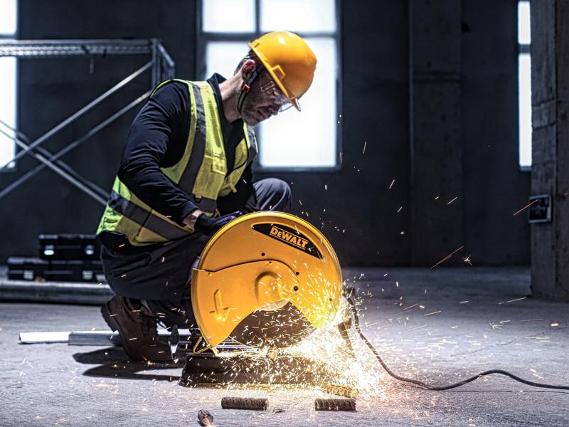 A worker wearing a yellow hard hat and safety vest uses a yellow DEWALT D28730 cutting machine to cut metal pipes in an industrial setting, causing sparks to fly.