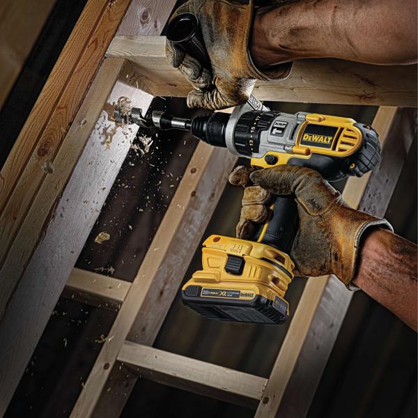 A person wearing work gloves uses a DEWALT cordless drill to bore a hole into a wooden beam, with wood chips flying as the drill operates.