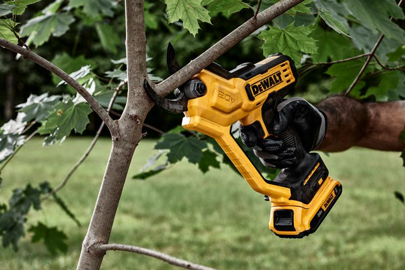 A person wearing a black glove is using a yellow DEWALT 20V cordless electric pruner to cut a branch from a tree outdoors.