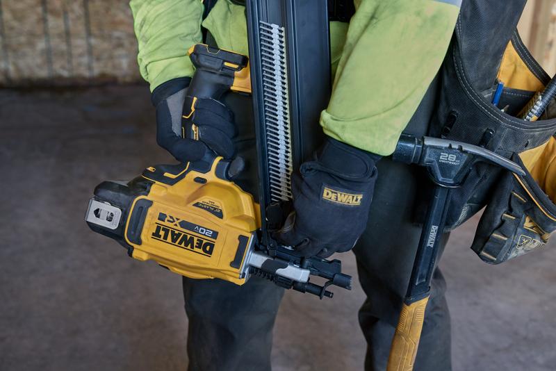 A person wearing gloves is holding a DEWALT DCN920B cordless framing nailer, with nails loaded into the magazine. The person is dressed in work attire and stands inside a construction site.