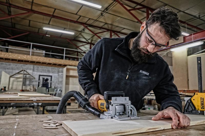 A person using a DEWALT DW682K joiner tool to work on a piece of wood in a workshop. The person's face is blurred out for privacy. The workspace includes tables, wood pieces, and visible workshop equipment.