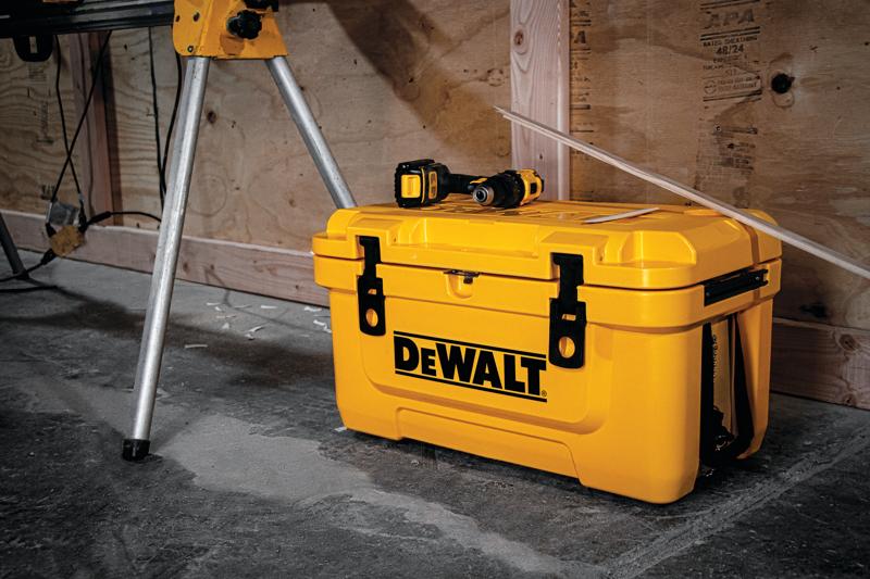 A yellow DEWALT branded toolbox with black latches is placed on a workshop floor. A cordless drill sits on top of the toolbox. The background features wooden walls and part of a metal tripod.