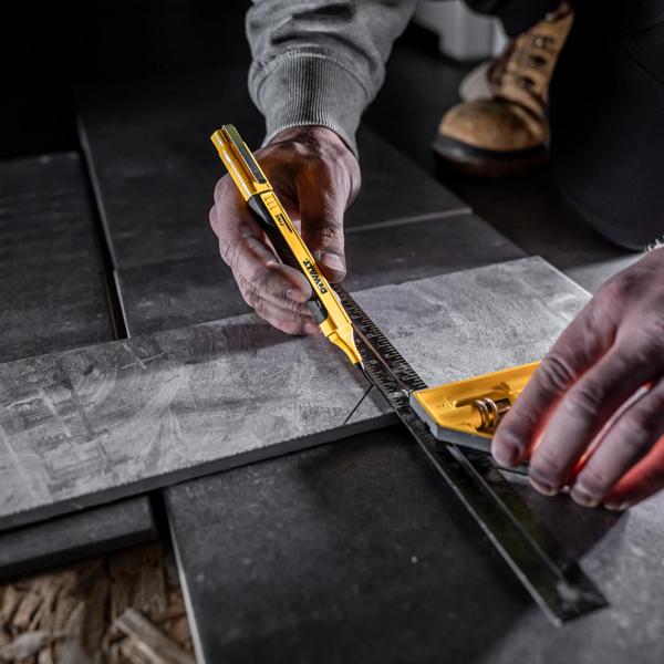 A person is using a DEWALT utility knife and a yellow measuring square to cut or mark a gray tile. The scene shows close-up hands working, with a ruler positioned for accurate measurement. The surface is a tiled floor, and the person is wearing work boots and a gray sleeve.