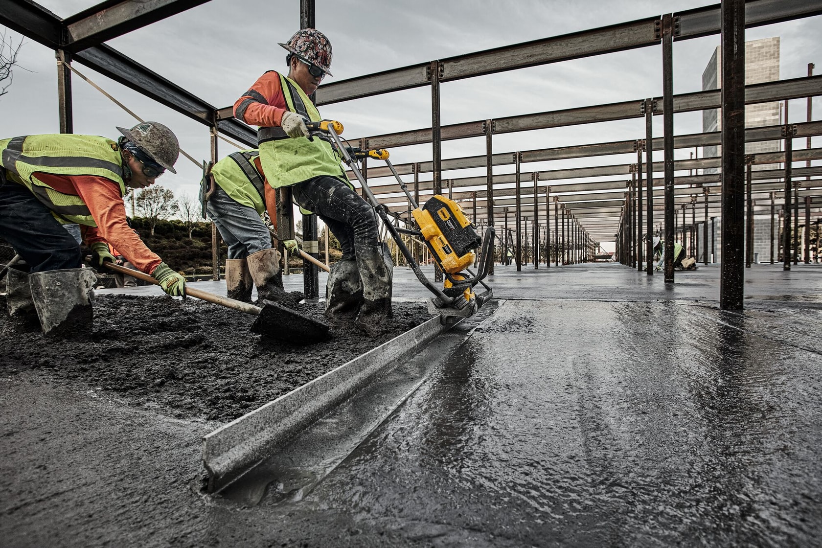 Construction workers in safety gear are using a DEWALT power tool to smooth freshly poured concrete on a large building site with steel beams and framework.