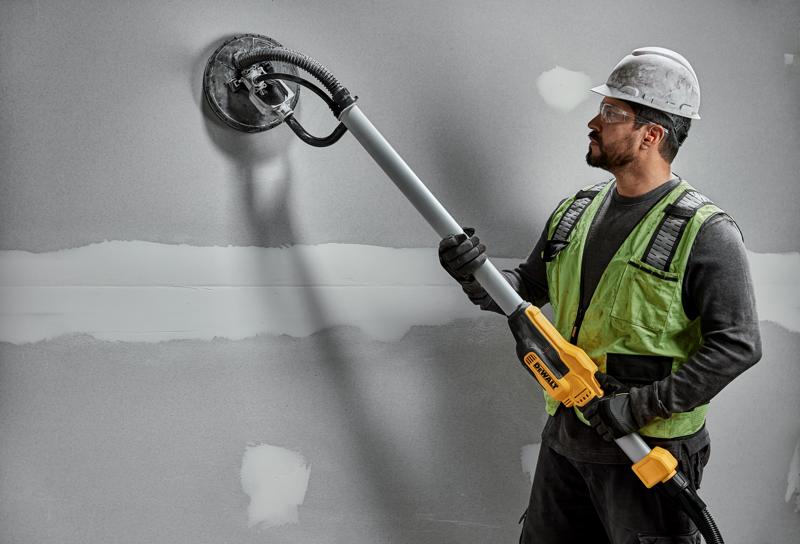 A construction worker wearing a hard hat and safety vest uses a DEWALT DWE7800 drywall sander with a telescopic handle to sand a gray wall.