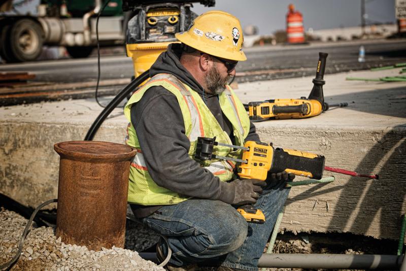 A construction worker wearing a yellow hard hat, safety vest, and gloves is using a DEWALT power tool to drill into concrete at an outdoor construction site. The worker's face is blurred for privacy. Other DEWALT tools and construction equipment are visible in the background.