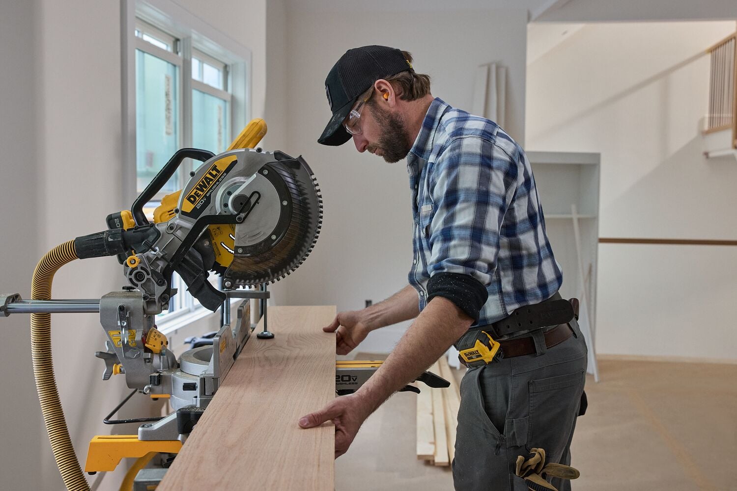 A person is using a DEWALT miter saw to cut a wooden board inside a workshop. The individual is wearing a plaid shirt and a black cap, and their face is blurred for privacy.