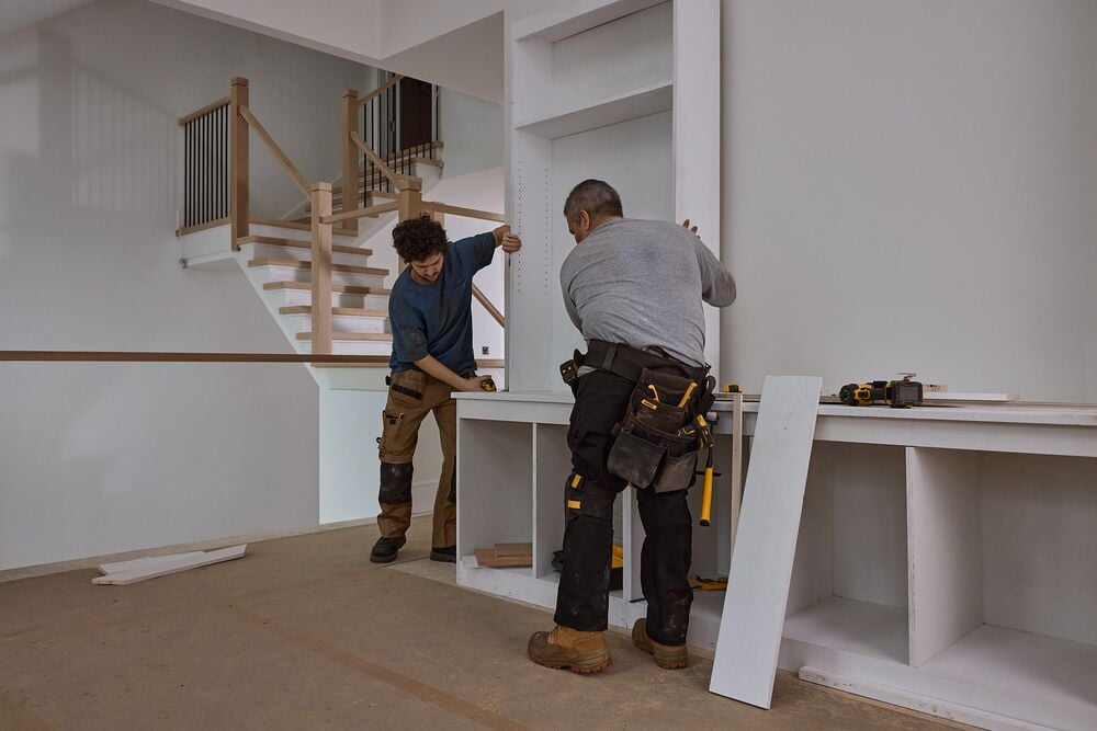 Two people working on assembling white shelving units in a home interior. One person uses a tool while the other holds a vertical panel. Both are wearing work clothes and tool belts. DEWALT tools and materials are visible on the countertop. In the background, there is a staircase and unfinished flooring.