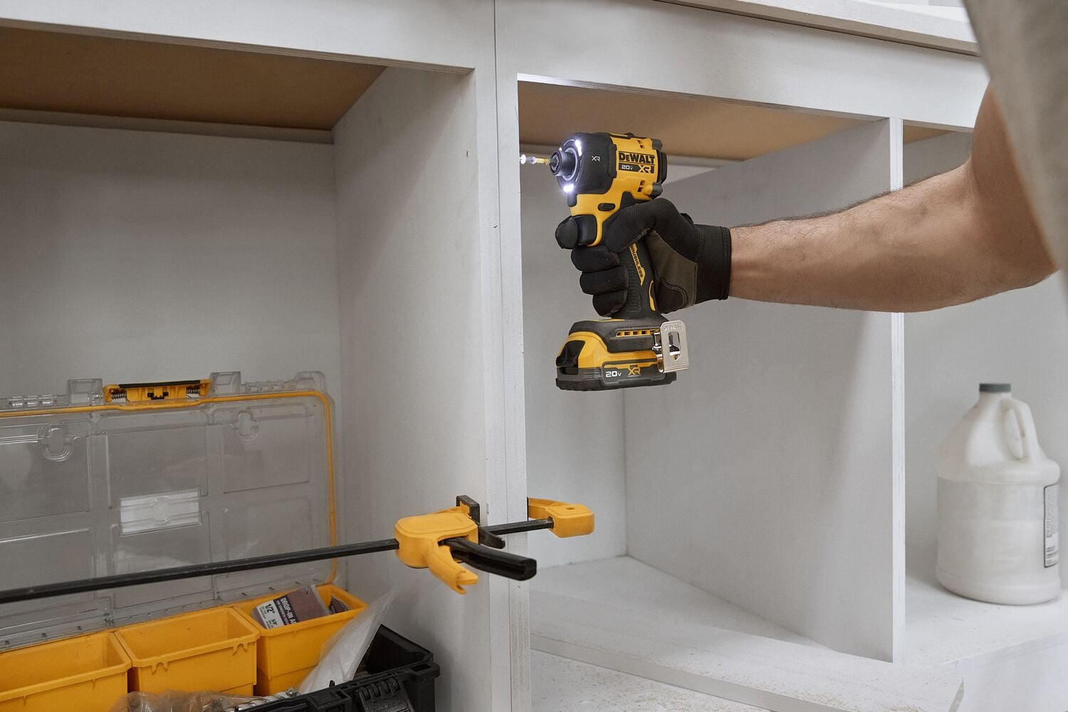 A person wearing a black glove is using a DEWALT cordless impact driver to drill into a white cabinet. A yellow clamp holds the cabinet in place and a yellow tool organizer box is visible nearby.