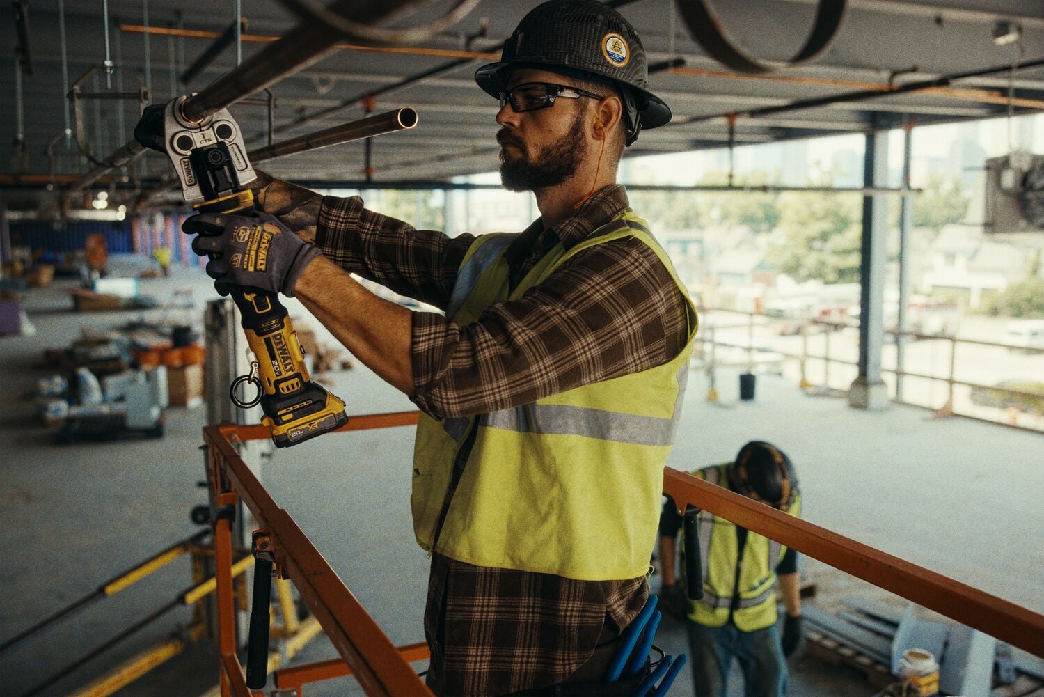 A person wearing a safety vest, gloves, and a hard hat is using a DEWALT DCE210D2 cordless tool to work on a pipe in a construction setting. The person is standing on an elevated platform and appears to be performing plumbing or pipe maintenance. The face of the person is intentionally blurred for privacy.