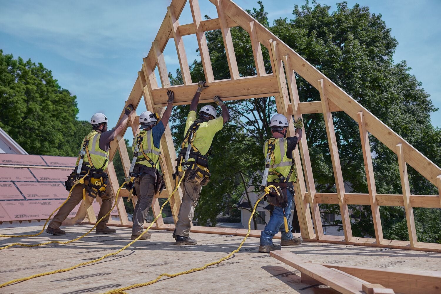 Four construction workers wearing safety gear and harnesses are lifting and positioning a wooden roof frame on a building site. Trees and clear sky are visible in the background.