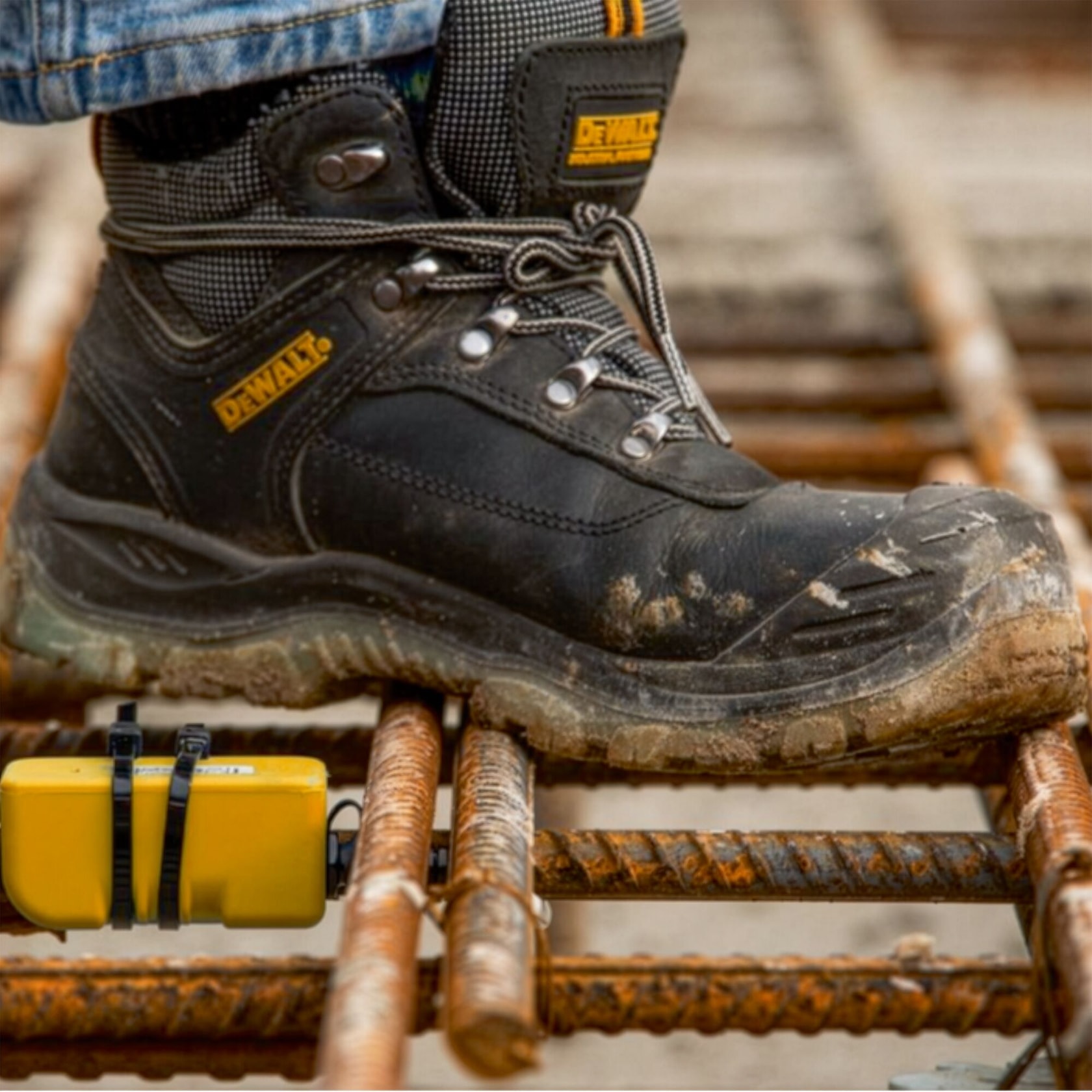 A close-up of a muddy DEWALT safety boot stepping on a rusty steel rebar grid at a construction site. A yellow electronic device is attached to the rebar below the boot.