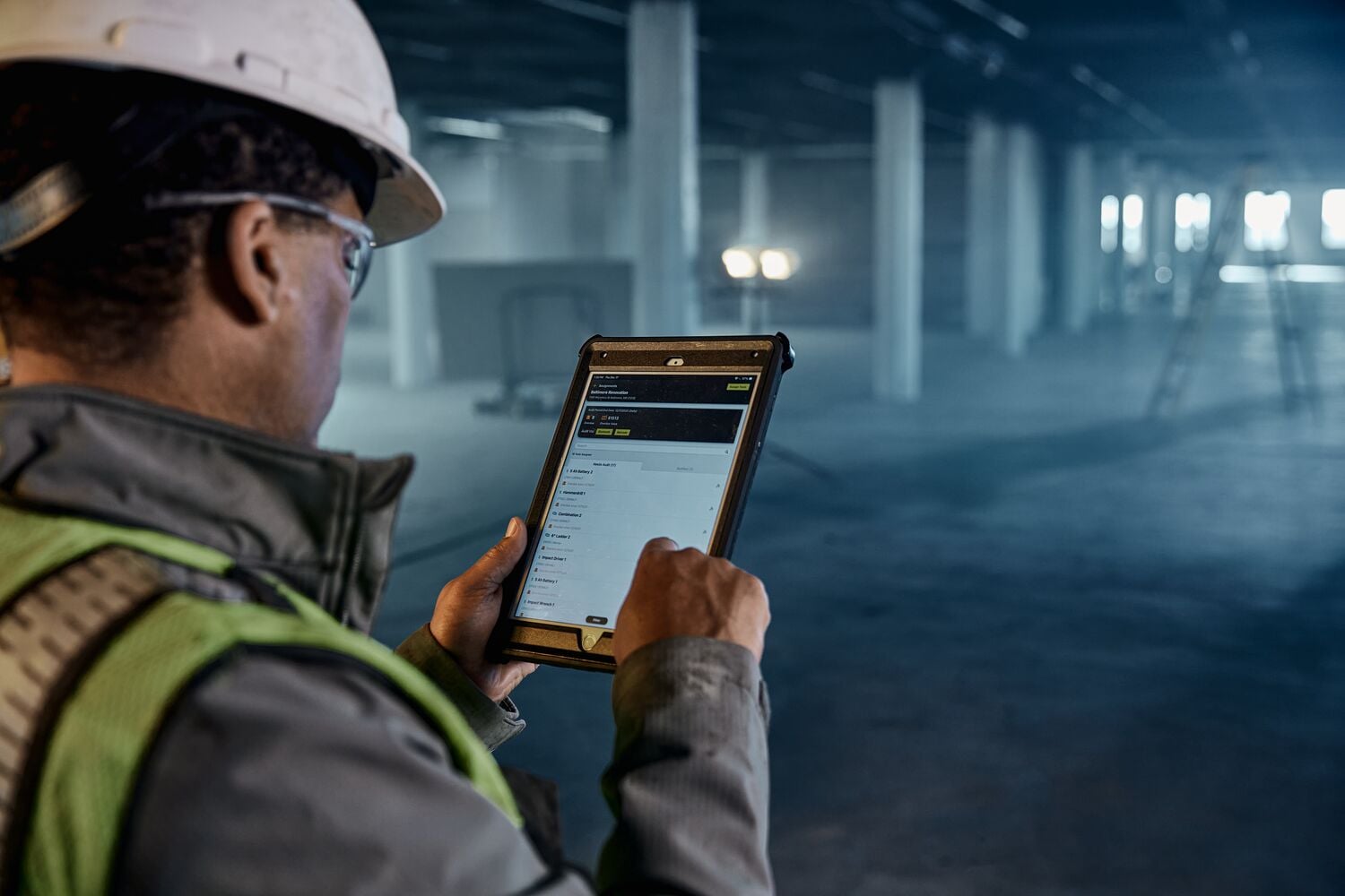 A person wearing a safety helmet and vest is using a tablet device in an industrial or construction setting. The screen displays a digital application interface. The environment is spacious with concrete pillars and construction equipment.