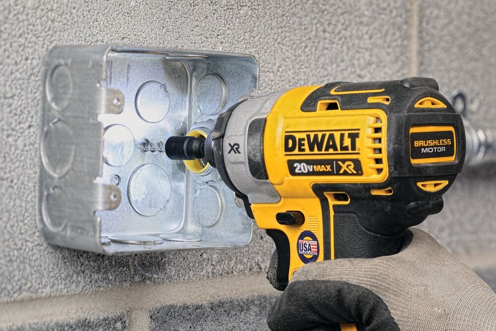 A DEWALT 20V MAX XR brushless motor impact driver being used to secure screws into a metal electrical box mounted on a concrete wall. The tool is held by a gloved hand.