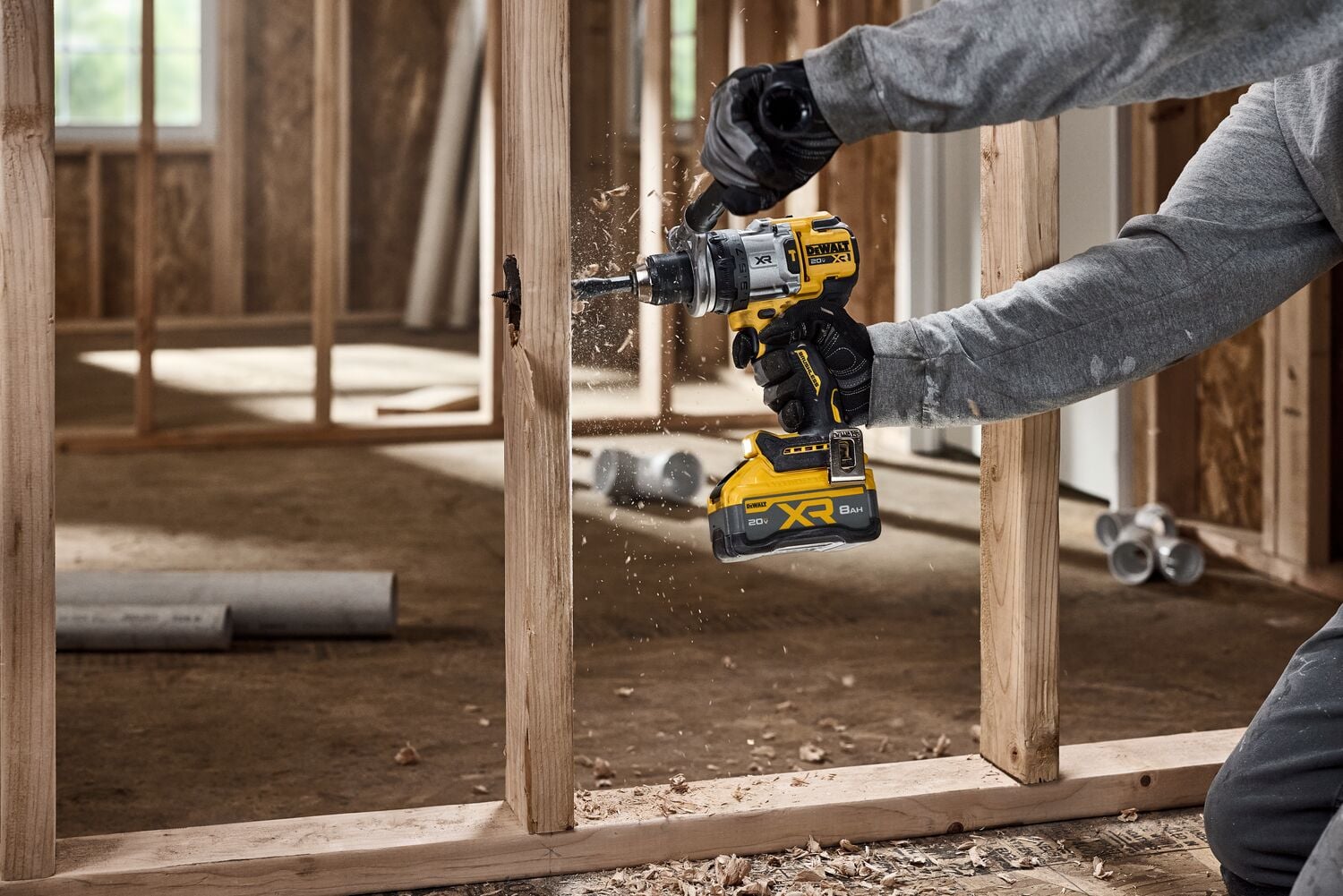 A person using a DEWALT cordless drill to bore a hole into a wooden stud in a construction setting. Wood shavings are visible as the drill operates, and the person is wearing gloves and a long-sleeve shirt.