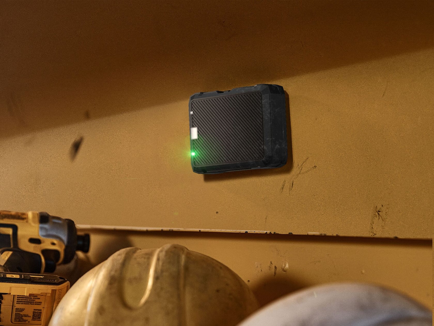 A DEWALT wireless tool tracker with a green indicator light is attached to a yellow metal surface. Below the tracker, a DEWALT power tool and construction helmets are partially visible.