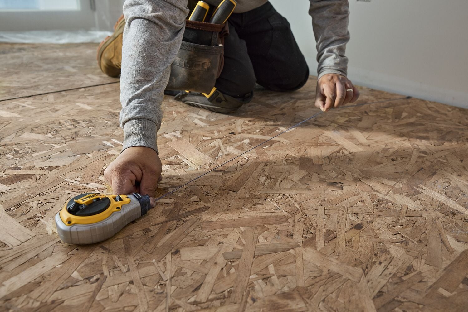 A person using a DEWALT measuring tape to measure wood flooring while kneeling, with a tool belt visible.