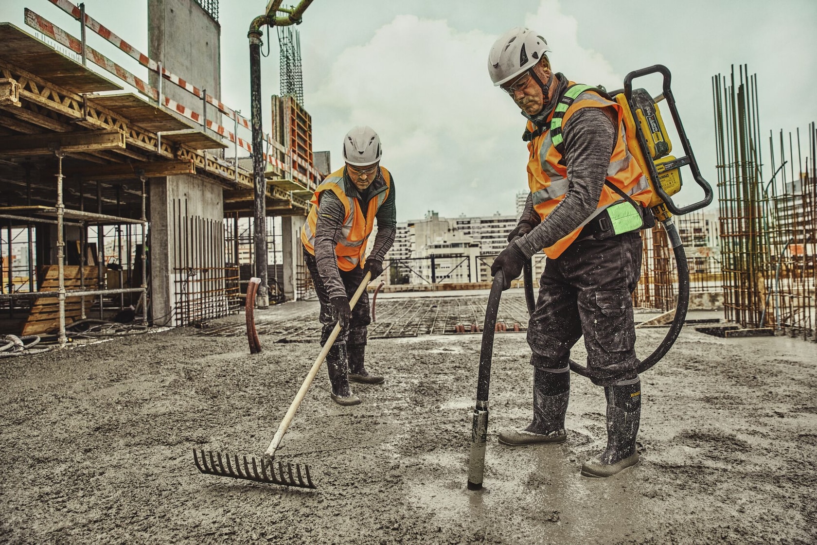 Two construction workers wearing safety helmets, gloves, and orange vests are working on a building site. One worker is holding a rake to level concrete, while the other uses a DEWALT backpack concrete vibrator. The background shows a partially built structure with steel reinforcements.