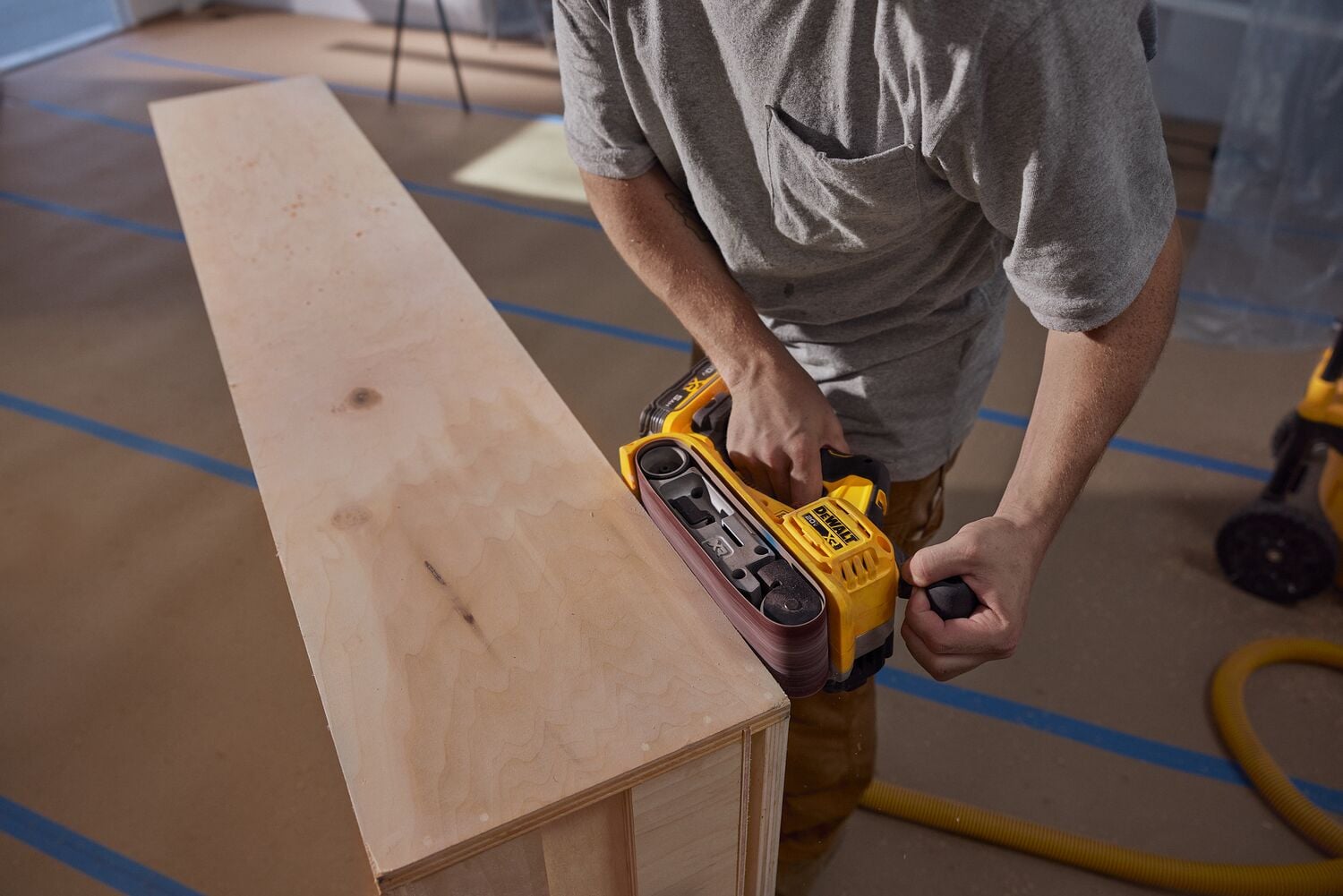 A person using a DEWALT belt sander to smooth the edge of a wooden board in a workshop setting.